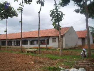 A photograph of a long, low whitewashed school building with an orange clay-tile roof, set among young saplings on bare reddish earth. A blue tarpaulin is stretched over part of the verandah. A bare-headed... no, a man wearing a white head cloth and white clothes crouches at the far right beside a stone at the edge of the plot. A stone bench sits on the grass in front, with a wooden door and window visible on the building. Overcast sky. A faint purple logo appears at the upper left and copyright text at the lower right.