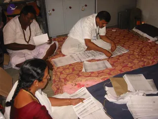A photograph of three people sorting printed paper sheets in a small storage room. A man in white kurta and white dhoti sits on the left, wearing a rudraksha mala and reading a sheet. A man in a white shirt sits on a red and gold patterned mat, sorting a stack of papers. In the lower left, a woman in a maroon blouse and white sari with a long braid and red bangles sorts printed pages. Behind them stand grey metal cabinets, a cardboard box and a dark bag. Piles of white papers lie on a blue cloth at right.