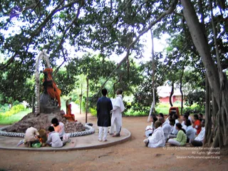Outdoor photo beneath large spreading banyan trees with aerial roots. A dark stone statue of a seated figure in orange-saffron cloth stands on a mound of earth inside a circular white-edged platform. Near it, a person in orange clothing sits by a small fire with offerings, and another person in white sits beside them. Two standing men are at the platform's edge: one in a dark blue tunic and white trousers, the other in an all-white kurta. About a dozen seated people in white and light clothes sit on the sandy ground to the right, and a red-roofed building shows in the background.
