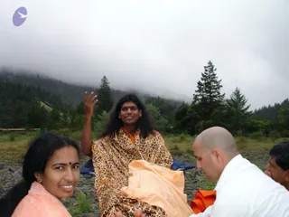 Outdoor photo on a rocky riverbank beneath misty, forested hills and tall conifers. A man with long, swept-back dark hair, wearing a leopard-print shawl over orange clothing, sits in a blue folding chair, one hand raised, and appears to be speaking. A young woman in a peach salwar with a red bindi smiles at the camera in the foreground. A bald man in a white shirt leans in at lower right beside an orange cloth, and another man is partly visible at the right edge. a person: the seated figure has long, swept-back hair and a leopard-print shawl over orange clothing, consistent with the page's a person reference.