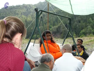 Outdoor gathering beside a river in a forested valley. A man with long dark hair and a red tilak on his forehead sits on a green metal swing frame under a green canopy, wearing a bright orange puffer jacket and with a white cloth draped over his lap. Around him, several people sit on the ground and chairs: a woman in the foreground with a ponytail and red knit sweater, a bald man in a white shirt, an older man with glasses in an olive jacket, and two men in the background, one in dark clothing and one in a yellow-collared top. Pine-covered hills and gravel riverbank fill the background.