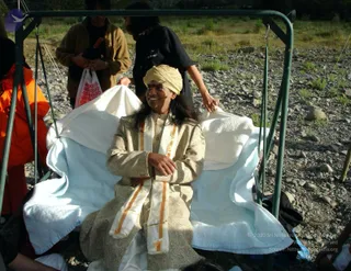 A man sits on a light blue and white towel-draped swing frame set on a rocky riverbed with dry grass behind. He wears a pale gold brocade kurta-style outfit with a cream and orange-bordered shawl draped over his shoulders and a pale yellow turban, smiling and looking to the side. Behind him, a person in a black top with a white logo leans in to adjust the towel, and another woman in a brown jacket holds a white bag with red print. A person in an orange jacket stands at the left edge. Appearance-based cues including the turban, long hair, gold brocade and shawl are consistent with a person seated on the swing, but the image is too uncertain to confirm.