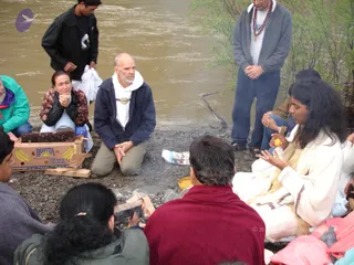 A photograph of a riverside homa on a pebbled bank beside a brown, fast-moving river, with a small fire of split wood burning on the ground. A long-haired person in a white robe with brown trim sits cross-legged at right, holding a yellow fruit and gesturing toward the fire. Opposite them, a person in a maroon shawl sits with their back to the camera. Around the fire, several devotees sit or kneel: a man in a navy jacket with a white hooded top kneels with hands folded; a woman in a floral jacket holds her hands together near her chin; a person in a green jacket sits at left. Standing behind, a man in a grey sweatshirt wears a rudraksha mala and a white shirt, hands clasped, and another man in a dark jacket holds a white bag. A cardboard Bonita banana box and a plastic bag rest on the stones. Two people are partly visible at the lower left.
