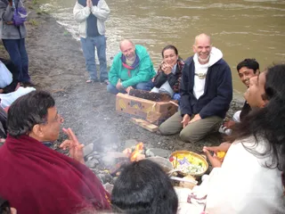 Outdoor riverside homa beside a muddy river bank with a small fire of logs and stones. Around the fire sit several people: on the left a man in a maroon shawl and glasses gestures with raised hands; a bald man in a teal-green jacket and purple scarf smiles; a woman with dark hair in a patterned shawl sits beside a bald man in a navy jacket with a white hooded top, who also smiles. A young man in a dark jacket smiles at the right edge. In the foreground a brass bowl holds flowers, fruit and offering items, and a cardboard Bonita banana box lies near the fire. Two standing people in jackets stand on the shore behind, one with hands pressed together in namaskar.