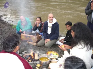 A riverside homa on a gravel bank beside a brown, fast-flowing river. A small fire burns on stones and split wood, with a steel pot of yellow liquid set near the flames and a brass tray of flowers and offerings nearby. A man in a teal jacket and a woman with dark hair, hands in anjali, kneel behind a cardboard Bonita banana box holding smoke. A bald man with a white beard in a navy jacket and white hood sits with hands folded. A young man in a black jacket watches. In the foreground, a figure with long dark hair in white robes holds a yellow flower and coconut-like offerings, and another person in a maroon shawl sits at the left edge.