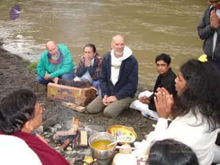 Outdoor photo on a pebbled riverbank beside muddy brown river water. A small wood fire burns on the stones in front of a steel pot of yellow liquid, a brass bowl of flowers and offerings, and a coconut. Several people sit or kneel in prayer with palms pressed together: a man in a green jacket, a woman in a floral top, a bald bearded man in a navy jacket and white hood, a young man in dark clothing, and a long-haired person in white in the foreground at right with a yellow flower in the hair. A man in a maroon shawl sits at left near the fire. A cardboard Bonita banana box and wooden planks sit on the bank.