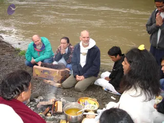 A photograph of an outdoor fire ritual on a rocky riverbank beside muddy brown water. A small fire of wooden logs burns in the foreground, with a steel pot of yellow liquid and a brass bowl of flowers and offerings beside it, and coconut halves on the ground. Around the fire, several people sit on stones and cloth with hands pressed in anjali. A man in a green jacket, a woman in a patterned top, and a bald bearded man in a navy jacket and white hooded top kneel with eyes closed. A younger man in a dark jacket sits nearby. On the right, a person with long dark hair in a white garment sits near a yellow flower in the hair, and a standing man in a grey jacket also holds his hands together. A cardboard box printed with Bonita banana branding sits beside the fire. This appears to be a homa held outdoors, with several participants seated in prayer.