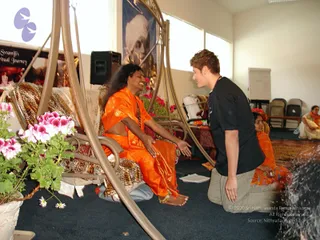 A photograph of an indoor teachers' training session in a bright hall with pale walls, blue carpet and folding chairs. A long-haired man in an orange-saffron silk garment sits on an ornate metal swing-style seat with leopard-print cushions and pink flowers beside him. A young man with short light-brown hair, wearing a black t-shirt, grey trousers and a beaded necklace, kneels before him with his hands extended in a gesture of reception. Behind them a woman in orange sits on a mat, and a poster reading 'Swamiji's Spiritual Journey' hangs on the wall with a portrait of a turbaned figure above. a person candidate is present, identified by long swept-back hair and saffron clothing with a rudraksha mala, though appearance is only partly clear.