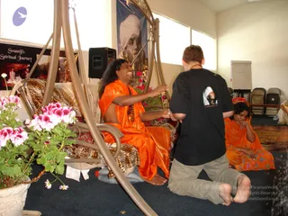 A photograph in a bright hall with cream walls, a white board and folding chairs. A man with long dark hair and an orange draped garment sits on a swing-style seat with leopard-print cushions, smiling and reaching to hold the hand of a kneeling man in a black t-shirt with a printed portrait and khaki shorts. A woman in an orange sari sits on the floor to the right, her hand at her face. Pink geraniums sit in a white pot at left, and a portrait of a turbaned figure is displayed on a blue banner behind the seat. A poster labelled "Swamiji's Spiritual Journey" hangs on the left wall. Two people are visible, and the seated man in orange is a person by his long swept-back hair, orange draping and his seated position on the decorated seat.