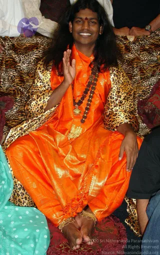 A seated man with long, dark, loose hair sits on a leopard-print cushioned seat, smiling and raising one hand in greeting. He wears a bright orange silk garment with gold zari patterns, a dark rudraksha mala with a gold pendant, and a gold ceremonial pendant at the chest. A red bindi marks his forehead and he wears gold anklets on bare feet resting on a red patterned cushion. Behind him are other people's sleeves, and a teal fabric edge appears at the left. Appearance-based a person: long swept hair, saffron-orange silk, rudraksha mala and gold ornaments, seated in a ceremonial manner.