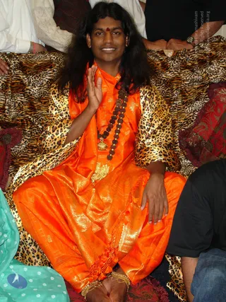 A seated man with long, dark, swept-back hair and a red bindi wears an orange silk garment with gold brocade and a leopard-print shawl draped over his shoulders. He raises one hand in a blessing gesture and wears layered rudraksha malas with a gold pendant. Gold anklets are visible at his feet. He sits on a leopard-print cloth over a red patterned cushion. Partial figures of other people in white and dark clothing stand or sit at the edges. a person: the orange robe, long hair, rudraksha malas and gold ornaments match the appearance cues, and the page does not state he is present at this event, so the label stays tentative.