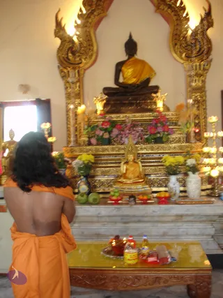 A photograph inside a Buddhist temple hall. In the foreground, a person seen from behind with long dark wavy hair, wearing an orange-saffron cloth draped over the shoulders and body, stands before an altar. The altar holds a large dark seated Buddha statue draped in orange-gold cloth, set within an ornate gilded arched frame with carved naga-style motifs. Below it sits a smaller gold seated Buddha image on a tiered gilded base, surrounded by pink, magenta and yellow flowers in vases, green coconuts, red-rimmed offering bowls of fruit, and tall candle holders with lit flames. On a low carved wooden table in front, a tray holds bottled water, a red-capped bottle, sweets and other small offerings. A gold Buddha statue is visible at the left edge near a window.