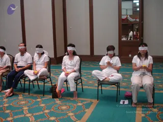Indoor hall with teal patterned carpet and cream walls. Several seated participants wear white clothing and white blindfolds, sitting on folding chairs with hands held together or holding yellow flowers. Some sit cross-legged on chairs and others are barefoot. A small figure in white stands in a mirror reflection at the upper right, beside draped red and white fabric and a decorative red display. Brown wooden paneling frames the room and a small offering lies on the floor.