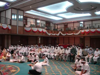A photograph of a large hotel ballroom with teal patterned carpet and recessed ceiling lights, where a seated group of about forty people, mostly in white clothing, sit on the floor and on chairs with white cloth blindfolds tied over their eyes. Several people in the front hold small objects in their hands. A balcony railing above is draped with red and white bunting and red rosettes, and a red-draped table with seated figures is visible at the far end of the hall.