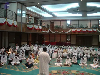 A large hotel ballroom with a teal patterned carpet and a balcony railing with red, white and gold draped bunting. Many seated participants wear white clothes and white blindfolds tied over their eyes, sitting cross-legged or on chairs in rows. In the foreground a man in white kurta stands facing the audience with his back to the camera, apparently leading the session. Several people in white and a few in yellow and orange sit in the middle rows. A red-draped table stands at the far end of the hall beside a doorway.