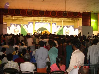 A photograph taken from the back of a crowded indoor hall, looking toward a lit stage with green and white decorative panels, golden pillars and a yellow circular dais. Many audience members stand and sit in rows of plastic chairs, several wearing white or striped shirts, a teal sari and a pink top. On stage a few figures in white clothing are visible near the centre, including one in white at the dais, with a camera on a tripod at the side. Ceiling fans and a patterned red and gold banner run across the top of the hall.