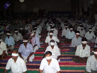 A photograph of a large indoor hall filled with seated men on colourful striped floor mats, most wearing white shirts and white or light dhotis and blindfolded with white cloth strips. Several participants wear name badges on their shirts. A man in the front centre has an orange and white shawl draped over his shoulders and a white cloth over his eyes. The hall has dim lighting, rows of chairs at the back, a large doorway, and a circular blue and white logo at the upper left. Faint copyright and source text appears over the lower right of the image.
