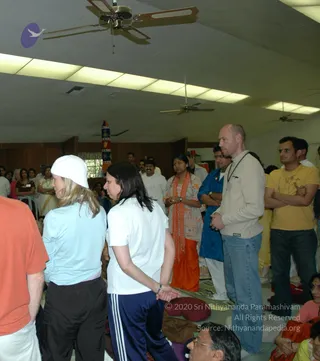 A photograph of a crowded indoor hall with a low ceiling, fluorescent panels and ceiling fans. In the foreground a woman in a white knit cap and light blue top stands beside a woman in a white t-shirt and navy track pants, both facing away from the camera. Centre, a woman in an orange and white shawl with an orange skirt stands with hands together. Beside her a man in a blue kurta with arms folded stands next to a bald man in a cream zip sweater and jeans. At right a man in a yellow t-shirt stands with arms folded. Several people stand or sit on floor cushions in the background, and a striped decorative pole stands at left.