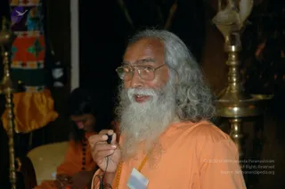 A bearded man with long grey hair and wire-rimmed glasses stands in the foreground, speaking and holding a small black microphone cord in his right hand. He wears an orange robe and a beaded amber-coloured mala, and a white name badge is pinned to his robe. Behind him, a second person in orange sits in soft focus beside tall brass oil lamps on stands, with a dark background and a colourful embroidered hanging at left.