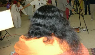 A photograph from an indoor hall with a bright studio light on the left and a tripod-mounted camera on the right. In the foreground, a person with long dark wavy hair and a saffron-orange garment is seen from behind, seated facing away. Behind them, a bald man in white clothes and a rudraksha mala bends forward with arms extended, and a person in a white t-shirt with a printed graphic stretches arms out, with others in white and colourful clothing nearby. Electrical cables and equipment lie on the floor.