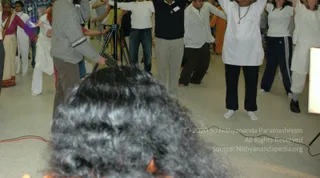 A photograph of a large indoor group in a tiled hall, with many participants standing with arms raised in a group movement or exercise. In the foreground, a blurred close-up of long dark curly hair fills the lower centre, with a tripod and camera on a stand behind it. A man in a grey knit sweater and khaki trousers holds a small device near the tripod. On the right, a person in a white tunic and black trousers stands barefoot with arms raised, wearing a long beaded necklace. Other participants wear white shirts and tracksuits, and some wear orange and saffron garments.