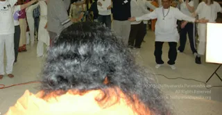 A photograph of an indoor gathering in white clothing with a blurred foreground of long dark curly hair above orange-lit flames. Behind it, a man in white kurta and dark trousers stands with arms outstretched beside a crowd of people in white and light clothing, some with hands raised. A tripod and a white panel are visible at the right edge, and a small badge hangs on one shirt. Floor and room details are plain.