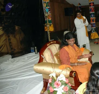 A photograph of a seated figure with long dark hair, wearing an orange-saffron shawl over a rust-brown top and a red bindi, sitting in an ornate gold-upholstered wooden throne with a gold cushioned armrest. The figure gestures with open hands. Beside the throne stands a man in white kurta and orange scarf with hands pressed together, and a brass oil lamp stands near the dark draped backdrop. A bouquet of pink gerbera, roses and white lilies rests at the base of the throne, and the front of the frame shows the back of another person's head in the corner. The wooden-paneled hall has colourful embroidered pillar covers in red, teal and orange. Two water bottles and a thermos sit on a white-draped table to the left. Printed photo credit reads Sri Nithyananda Paramashivam and