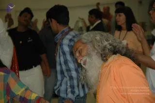 A photograph of an indoor gathering where a bearded elderly man with long grey hair, wearing an orange-saffron robe and an orange bead mala, leans forward with eyes closed while others around him raise their hands. Behind him stand several people in casual clothes, including a man in a blue plaid shirt with a red-brown shawl, a man in a black t-shirt, and a woman in a peach top with a necklace. A person in a colourful checked garment is partly visible at the lower left.