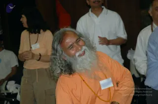 A photograph of an indoor gathering where an older man with long swept-back grey hair and a full white beard, wearing an orange-saffron robe and an orange-amber bead mala, smiles broadly with his head tilted. He wears a white name badge on his chest. Behind him, a woman in a peach top with a rudraksha-style red-brown bead necklace and tan cargo pants stands with hands clasped, and a man in a white polo shirt gestures. A man in a light blue shirt and a partly visible man in white stand at the right edge against a dark wooden wall.
