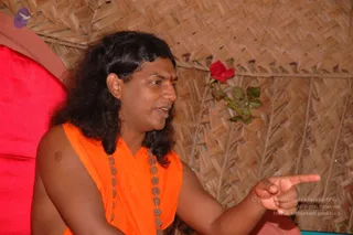 A man with long, swept-back dark hair and a red tilak mark on his forehead sits against a red cushion, wearing a sleeveless saffron-orange garment with a long beaded necklace of round medallions. His bare right arm is extended to point toward the right. Behind him is a woven bamboo or thatch wall with a red rose stuck into it. The setting appears to be a simple hut-like space.