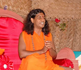 A man with long, swept-back dark hair sits on a red cushioned throne against a woven bamboo wall, speaking with his hands joined at chest level. He wears a sleeveless saffron-orange robe, a long rudraksha mala draped over his shoulders, and a red bindi on his forehead. A single red rose is tucked into the bamboo wall behind him, and red satin cushions and a pink arched backrest frame the seat. Swamiji (probable): the long swept-back hair, saffron robe and rudraksha mala match the appearance-based cues on this page, and the seated figure is addressing an audience.