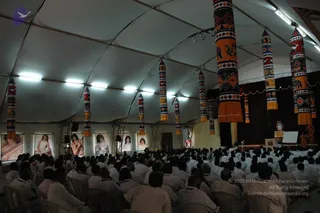 Interior of a large white tent-style hall where a dense audience of several hundred seated people, mostly dressed in white, faces a stage at the far end. Tall vertical cylindrical banners with bold geometric, floral and animal patterns in red, orange, yellow and black hang from the ceiling. Along the left wall, large framed portrait posters show a bare-shouldered figure with long hair and a seated figure. On the stage a figure in orange clothing sits beside a white board and a decorated pot. Bright ceiling lights and speakers are visible.