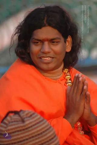 Close-up photo of a smiling man with long, wavy dark hair and a small red tilak on his forehead, wearing a bright orange shawl over a beige turtleneck. He holds his palms pressed together in anjali namaste, and a gold ornamental necklace and crystal-beaded mala hang at his chest. A striped knitted cap is partly visible in the bottom left corner. The background is a soft blur of green and blue.