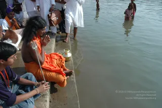 A photograph taken at a riverside ghat where a sannyasi in saffron-orange cloth sits on the stone steps with palms pressed together in namaste, a trident (trishul) resting beside him and a brass kalasha-style pot near his feet. He has long dark hair and bare shoulders. Beside him a young man in a purple shirt and jeans, wearing a beaded bracelet, sits with hands folded. Behind them devotees in white clothes stand at the water's edge, and several people wade into the river. The water is calm and brownish, and the scene suggests a ritual bathing or prayer at the river.