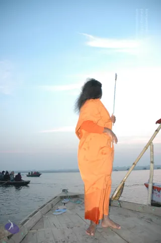 A barefoot person in a long saffron-orange robe stands at the front of a wooden boat on a wide river at dawn, holding a tall thin metal staff upright in one hand. Long dark hair falls loosely down the back. Behind, small wooden boats carry several seated passengers across pale blue water under a hazy sky. A bamboo oar leans on the right side of the boat, and a pair of blue flip-flops lies on the deck. The setting suggests a river crossing on the Ganga at Varanasi.