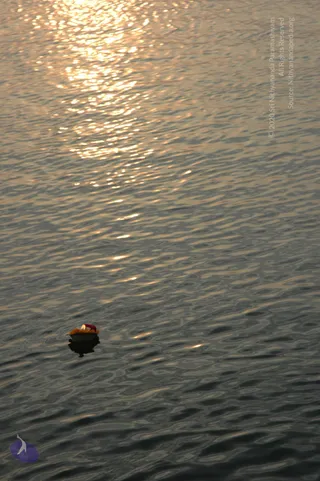 A small floating offering rests on rippling river water at sunset, with golden sunlight glinting across the surface. The offering is a small leaf or clay bowl holding orange marigold petals and a lit oil lamp or candle with a red element. Its reflection is visible beneath it. No people are visible in the frame.