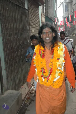 A photograph of a long-haired man in an orange saffron robe walking down a narrow street in Varanasi. He wears a heavy garland of orange and yellow marigolds with red flowers, and a red bindi on his forehead. Behind him a man in a blue jacket holds a mobile phone, and another man in white walks past. Red flags hang across the lane above, and shuttered shop fronts line the street.