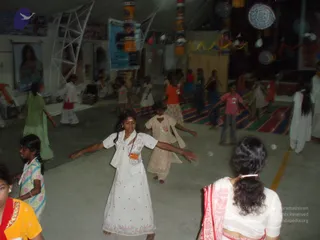 A photograph taken indoors at night of a large hall with a smooth concrete floor where children and young people are dancing or moving with outstretched arms. In the foreground a girl in a white dress with a white scarf and a small tilak on her forehead spreads her arms, while a girl in a beige salwar kameez with a dupatta and a girl in a pink top and jeans move nearby. A woman in a white blouse and pink-and-white sari stands with her back to the camera at lower right. Behind them, a girl in an orange top and others in white and green outfits dance on a striped carpet beneath hanging decorations, paper lanterns, streamers and a large blue banner. A few white-clad figures stand at the right edge.