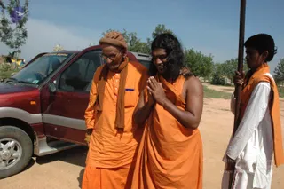 A photograph of three men on a dusty dirt road beside a dark red SUV. In the centre, a man with long, swept-back dark hair and a bare chest wears a saffron wrap and presses his palms together in añjali greeting while smiling. He has a distinctive appearance: long hair and a saffron wrap. An elderly man on the left wears saffron robes, a brown head wrap and a brown shawl, with glasses. On the right, a young man in white with a saffron shawl holds a long wooden staff. Trees and a blue sky are behind them.