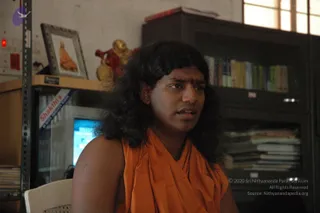 Photograph of a long-haired figure in a saffron-orange wrap seated indoors in an office-like room, speaking. A red-and-gold deity figurine sits on a shelf beside a framed photo of a saffron-robed figure, with a computer monitor, bookshelves and a stack of papers behind. The person wears a red forehead mark and a rudraksha-style pendant on the chest. a person appearance-based label: long swept-back dark hair and saffron clothing, with identity uncertain.