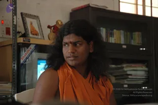A person with long, dark, wavy hair sits in an office-like room, wearing a saffron-orange shawl wrapped over one shoulder and a red bindi on the forehead. Behind them stand a wooden bookshelf filled with books and a tall dark cabinet with stacked papers. On the left, a framed picture of a saffron-robed figure rests on a shelf beside a small golden and red figurine, and a computer monitor is visible. The person looks off to the side with a serious expression. a person: the long hair and orange shawl are visible cues, so this is a candidate in person.