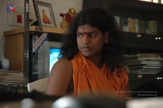 A photograph of a man with long, dark, swept-back hair and a small red tilak on his forehead, wearing a loose orange-saffron wrap over his shoulders and an upper body partly bare. He sits in an office or study room, looking off to the side, with a desk in the foreground. Behind him are a framed picture of a robed figure, a small golden figurine, a computer monitor, shelves of books and stacked papers. Appearance-based label: a person, given the long hair, saffron wrap and tilak cues; the page states he was not at this event, so this is an appearance-based candidate only.