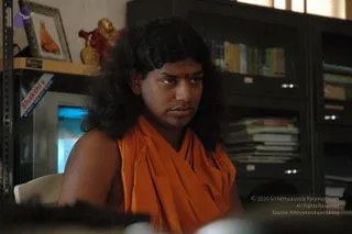 A photograph of a young man seated indoors in front of a metal office cabinet and bookshelves. He has long, dark, wavy hair and wears a loose orange saffron wrap over a bare shoulder, with a red bindi on his forehead. He looks off to the side, attentive. Behind him on a shelf sit a framed portrait of a saffron-clad man labelled as a printed photograph, a gold figurine and a small red figure, plus a stack of folders. A white plastic chair and a blurred foreground object are visible.
