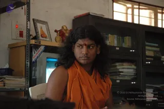 A photograph of a man seated in an office, wearing a loose saffron-orange wrap over bare shoulders, with long dark wavy hair and a red bindi on his forehead. He looks off to the side. Behind him stand a metal filing cabinet with books and folders, a computer monitor with a blue screen, and a framed picture of a saffron-robed figure on a shelf beside a small golden figurine. A red power indicator glows on the wall switch panel.