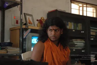 Indoor office photograph of a person with long, swept-back dark hair and a red tilak on the forehead, wearing a loose saffron-orange wrap over a sleeveless top. The person sits in a white plastic chair near a metal shelving unit and filing cabinets stacked with papers and books. Behind them on a shelf is a framed picture of a saffron-robed figure and a small golden figurine, with a computer monitor visible to the left. A tattoo is visible on the upper arm. The setting appears to be an administrative room.