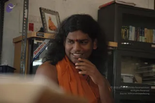 A photograph of a smiling man with long, dark, wavy hair and a small red bindi on his forehead, wearing an orange-saffron shawl draped over his shoulders. He rests his hand against his chin in a thoughtful pose. Behind him stands a metal shelving unit, a dark cabinet with books and folders, and a framed photograph on a shelf showing a figure in orange robes seated. A Shakthi's product box is visible at left. a person: the long swept-back hair, orange shawl and ornate styling match the page's album context, but appearance alone is uncertain.