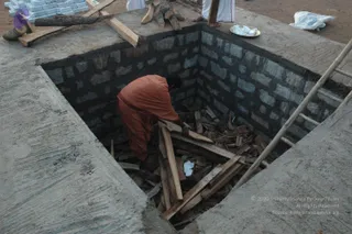 A photograph of a square concrete-lined fire pit under construction, with dark grey brick walls and a smooth concrete rim. A person in an orange-red robe with a dark sash is bent over inside the pit, arranging wooden logs and sticks. A bamboo ladder leans against the far rim, and a white-clothed figure stands at the top edge beside a plate of white offerings and stacked bricks. The sandy ground around the pit holds more wood and a white plastic bag.