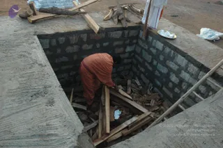A square brick-walled fire pit sunk into a concrete platform on bare ground. Inside the pit, a person in an orange-red garment bends low, arranging split wooden logs and kindling among scattered debris. A bamboo ladder leans against the far edge. Above, a white-clad figure with an orange cloth stands at the top edge beside a metal plate holding white items, with more stacked logs and a bundle of wood on the ground. Stacked white bricks sit at the upper left. The scene shows preparation of a fire pit for a homa, with no visible face or ceremony beyond the fire arrangement.