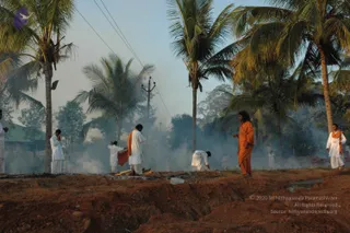 A smoky outdoor scene on reddish-brown earth beside tall coconut palms and a utility pole with wires. Smoke drifts across the ground where a fire pit has been prepared. A figure in a long flowing orange robe stands at the front right, long dark hair falling loose, with a hand raised near his chest. Several men in white dhotis and white shirts are scattered across the scene: one stands at left with hands folded in prayer, one at centre-left wears a white robe with an orange shawl over his shoulder and bends over the ground, another in white bends low near the fire, and a man in white with an orange shawl stands at the far right. Other people in white are faintly visible in the smoke. The setting appears to be a homa or yagna area being prepared outdoors.