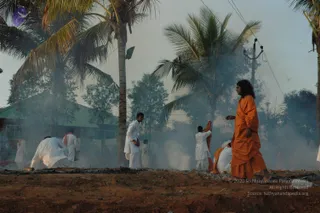 Outdoor fire ritual at dusk among coconut palms and a tin-roofed building, with smoke drifting across the scene. A man in a long flowing orange robe with long dark hair walks at right, gesturing with his hand. Several men in white dhotis and shirts kneel or stand at a smoldering fire pit with ash and burnt wood on red earth, one tending the fire and another lifting cloth. Electric wires and a utility pole stand in the background.